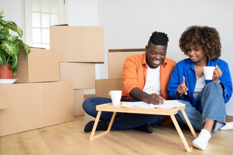 A young couple updating the moving checklist while surrounded by moving cartons in the concept of 'How to Create a Moving Checklist for Your Move to Lewisham'.